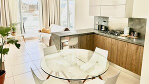 A modern kitchen with a central island, a dining area with a glass table, and a living space with a sofa and curtains. - Santa Venera - The Papillon Penthouse (L-Imsida)