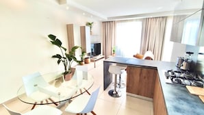 A modern kitchen with a central island, a dining area with a glass table and chairs, and a living area with a sofa and a TV. - Santa Venera - The Papillon Penthouse (L-Imsida)