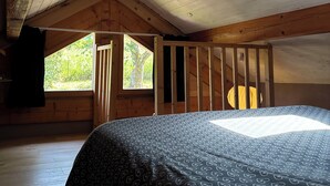 Ein Schlafzimmer mit schrägen Wänden, Holzbalken, einem Bett mit gemusterten Betttextilien und einem Fenster mit Blick ins Grüne.