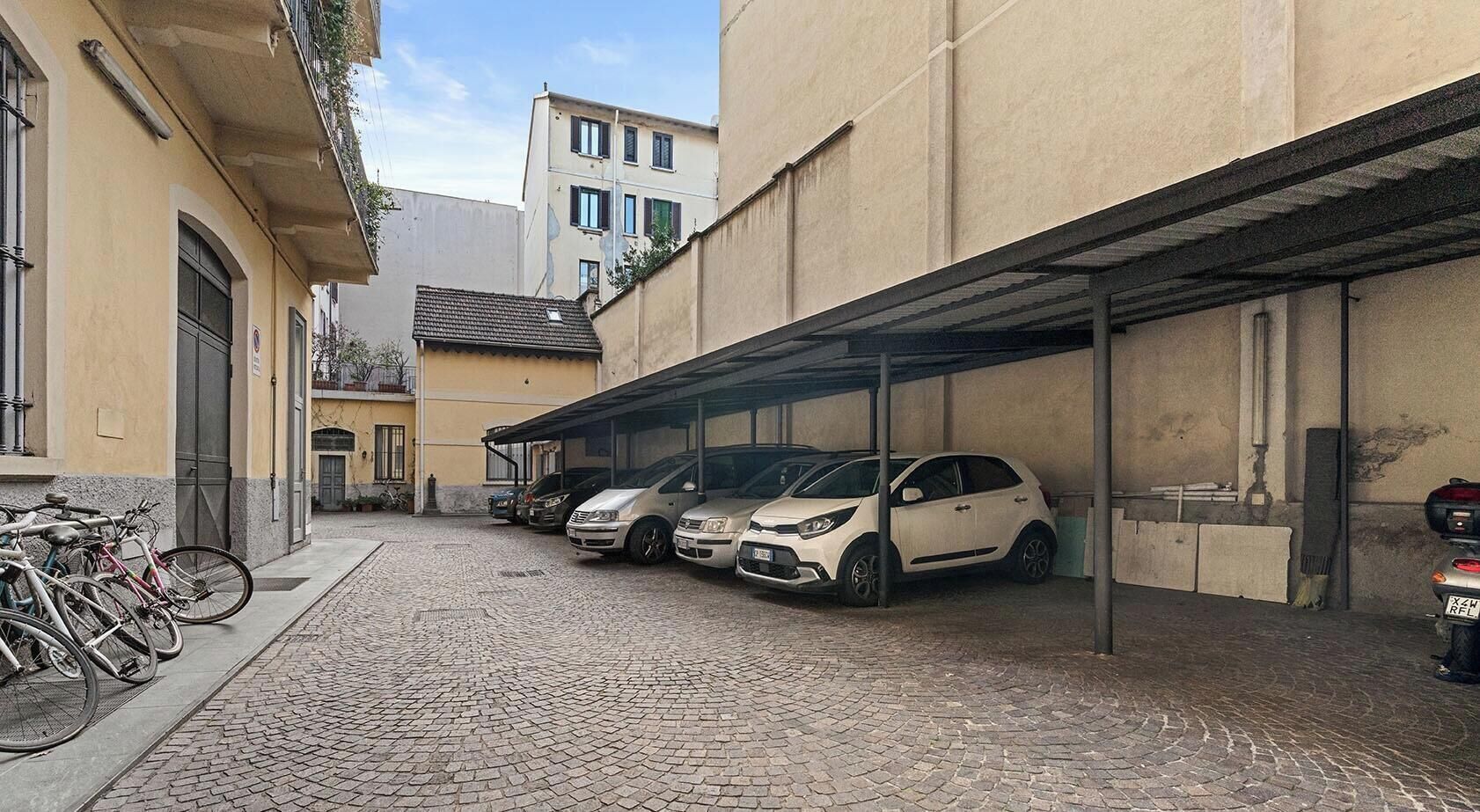 A parking area with several cars and bicycles, a cobblestone street, and buildings with balconies.