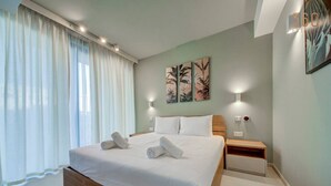 Ein ordentlich bezogenes Bett mit weißen Leinentüchern und zusammengerollten Handtüchern, ein Nachttisch mit Lampe und eine Wand mit drei gerahmten Kunstwerken.