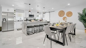 Une cuisine moderne avec des meubles blancs, un îlot en marbre et un coin repas comprenant une table et des chaises aux couleurs sombres.