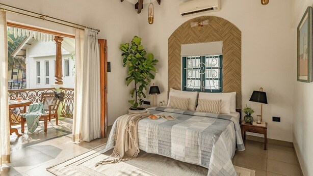 A bedroom with a large bed, a potted plant, a window with a herringbone pattern, and a balcony with a chair and table. - Sanctuary by RÊVE Escapes (Siolim)