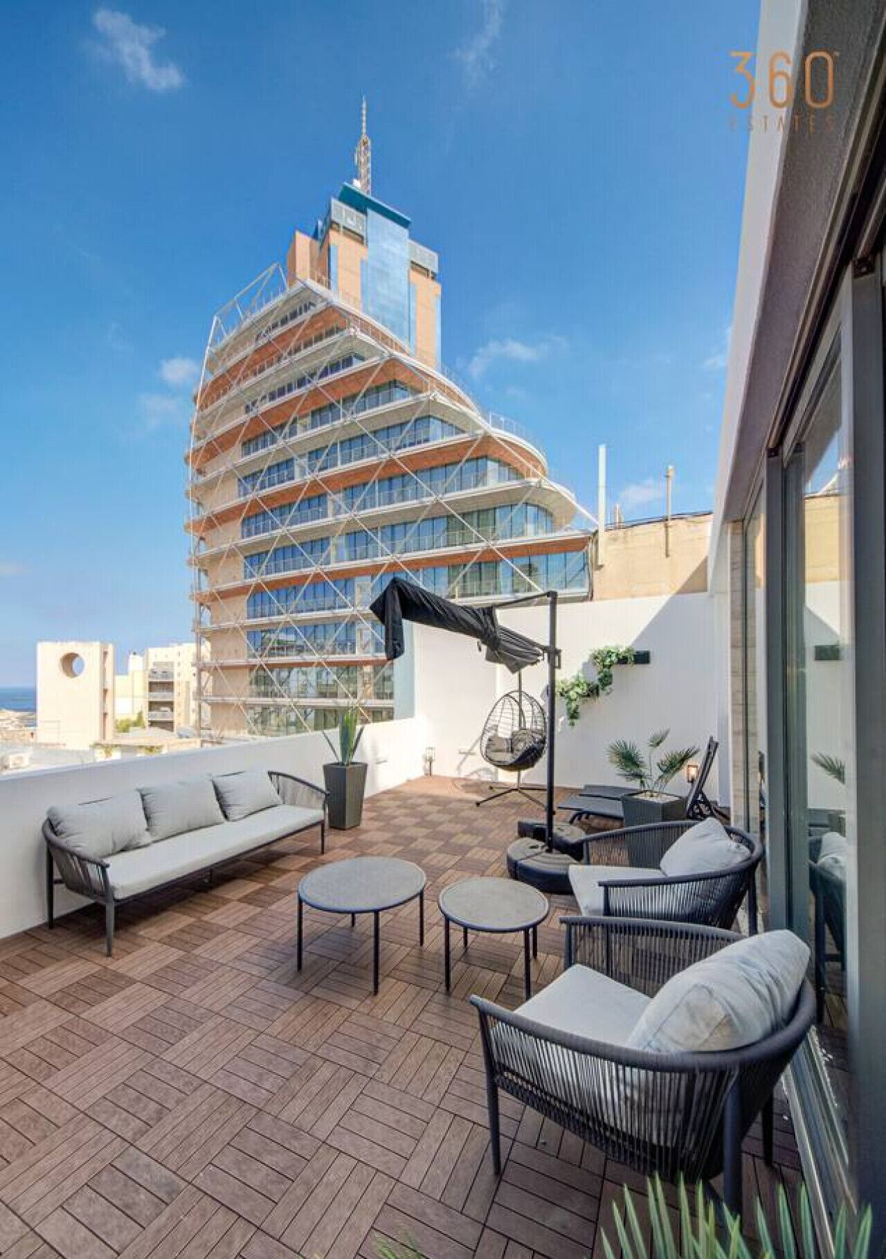 A rooftop terrace with outdoor furniture, a hanging chair, and a view of a modern building.