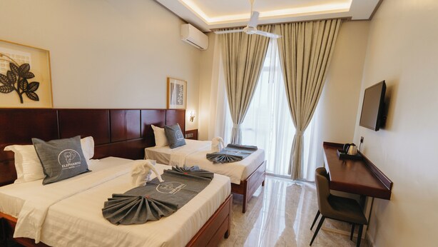 A hotel room with two beds, a desk, a chair, a TV, and a ceiling fan. - Elephantohotel (Zanzibar Town)