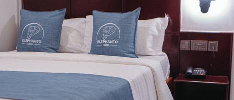 Una cama bien hecha con una colcha azul y dos almohadillas con el logotipo de un hotel, una obra de arte enmarcada sobre la cama y una mesita de noche con un teléfono y un objeto decorativo.