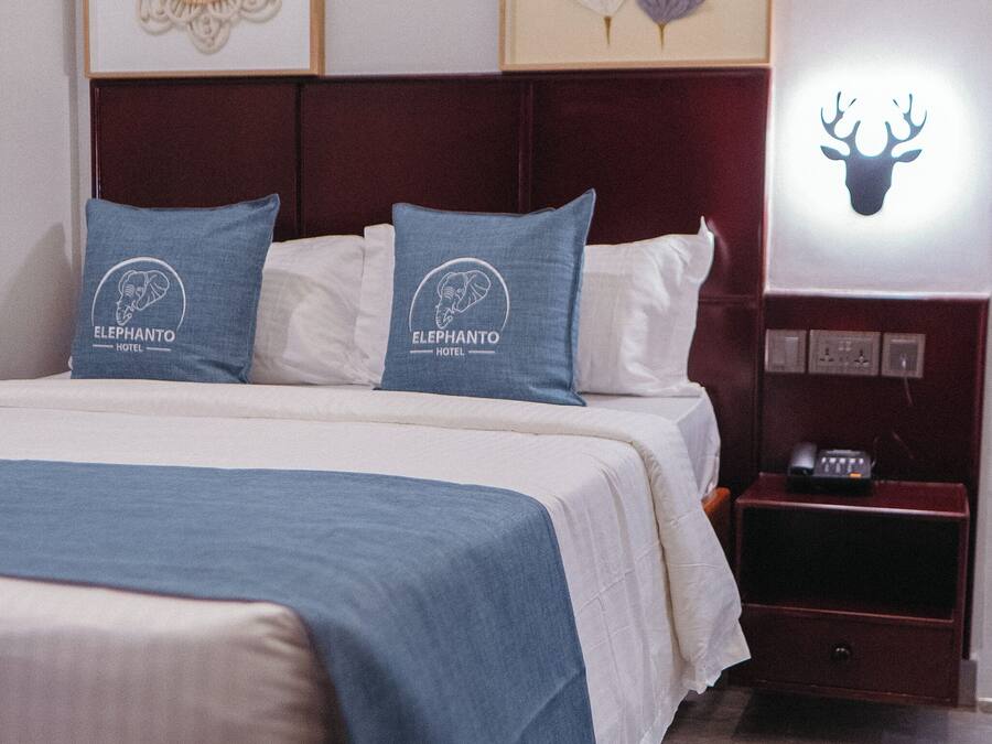Una cama bien hecha con una colcha azul y dos almohadillas con el logotipo de un hotel, una obra de arte enmarcada sobre la cama y una mesita de noche con un teléfono y un objeto decorativo.