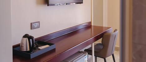 A hotel room with a wooden desk, a television mounted on the wall, a small refrigerator, and a trash can.