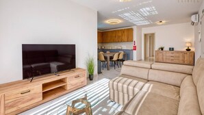 Un salon moderne doté d’un téléviseur à écran plat, d’un meuble de rangement en bois, d’un canapé beige et d’un coin repas avec une cuisine en arrière-plan.