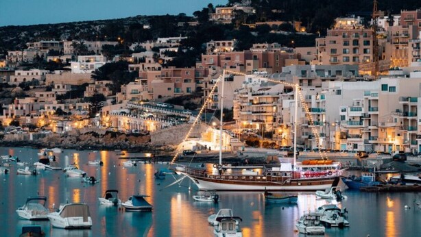 Une ville côtière au crépuscule, avec un port de plaisance rempli de bateaux, des bâtiments dotés de balcons et un grand navire amarré au centre.