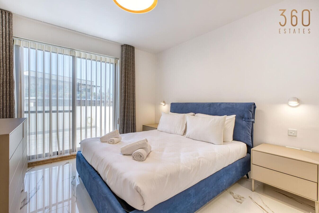 Una habitación de hotel moderna con una cama grande, una mesita de noche y un ventanal con cortinas.
