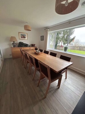 A dining room with a long wooden table, chairs, a lamp, and a window with a view of greenery.
