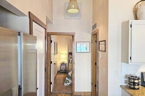 A hallway with a refrigerator, a kitchen area with cabinets, a hanging light fixture, and a framed picture on the wall. - Lakefront MM19|Top Floor|Cov Patio|Salt H2O Pool (Osage Beach)