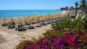 Una spiaggia con lettini e ombrelloni di paglia, una passerella in legno e vista sull'oceano e sugli edifici della città sullo sfondo.
