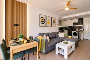 Un salon moderne comprenant un canapé, une table à manger et des chaises. Il y a également un coin cuisine équipé d’appareils électroménagers et doté d’un ventilateur de plafond.