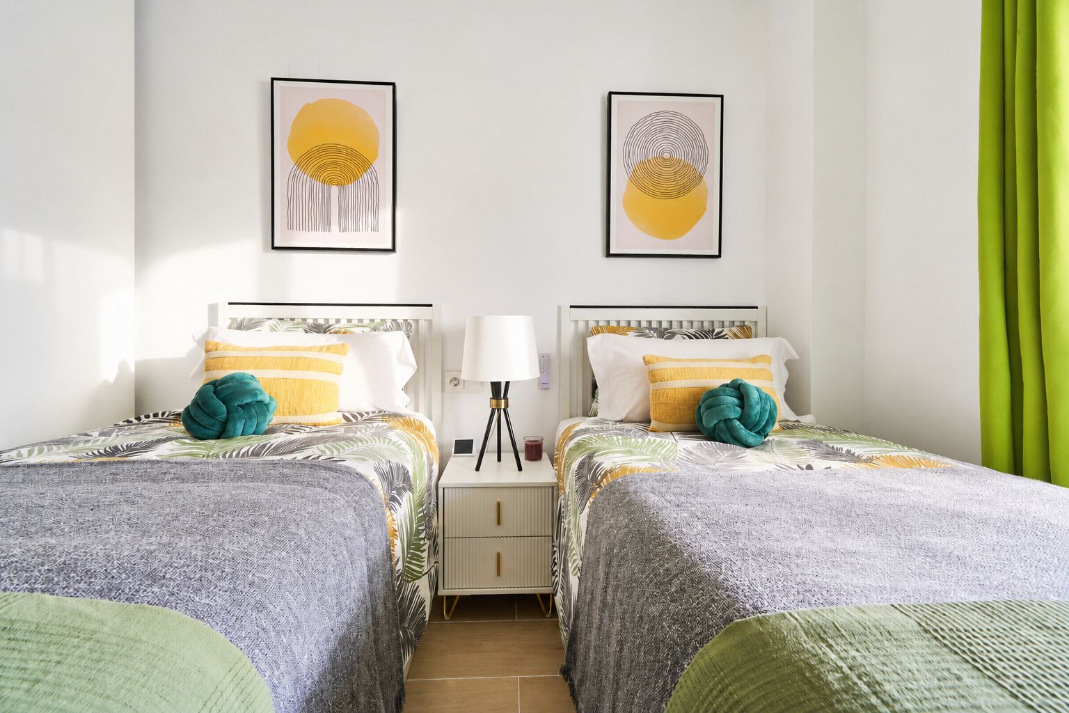 A hotel room with two beds, a nightstand, a lamp, and two framed artworks on the wall.