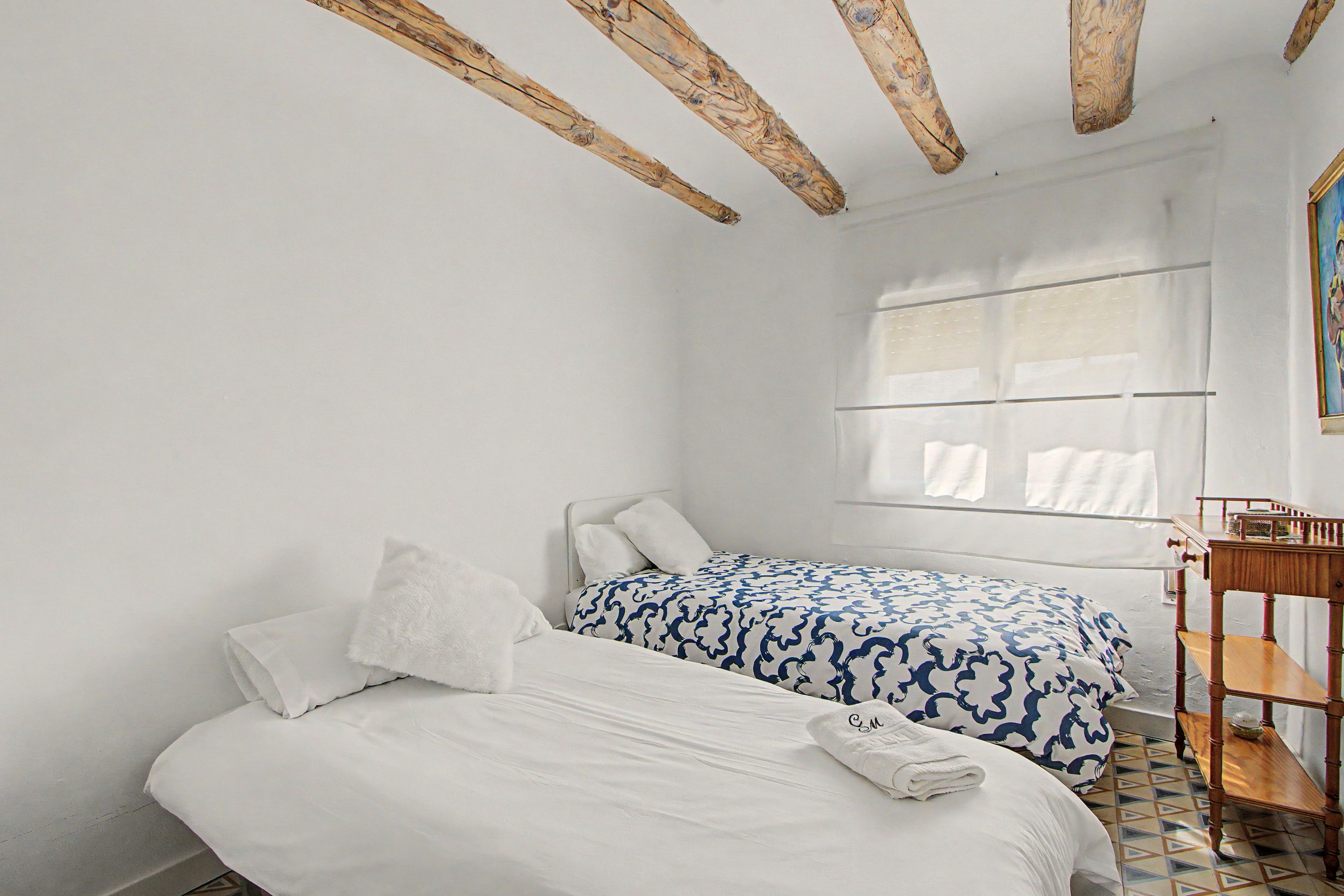Quarto com duas camas, uma janela com persianas, uma mesa de madeira e soalho em azulejos.