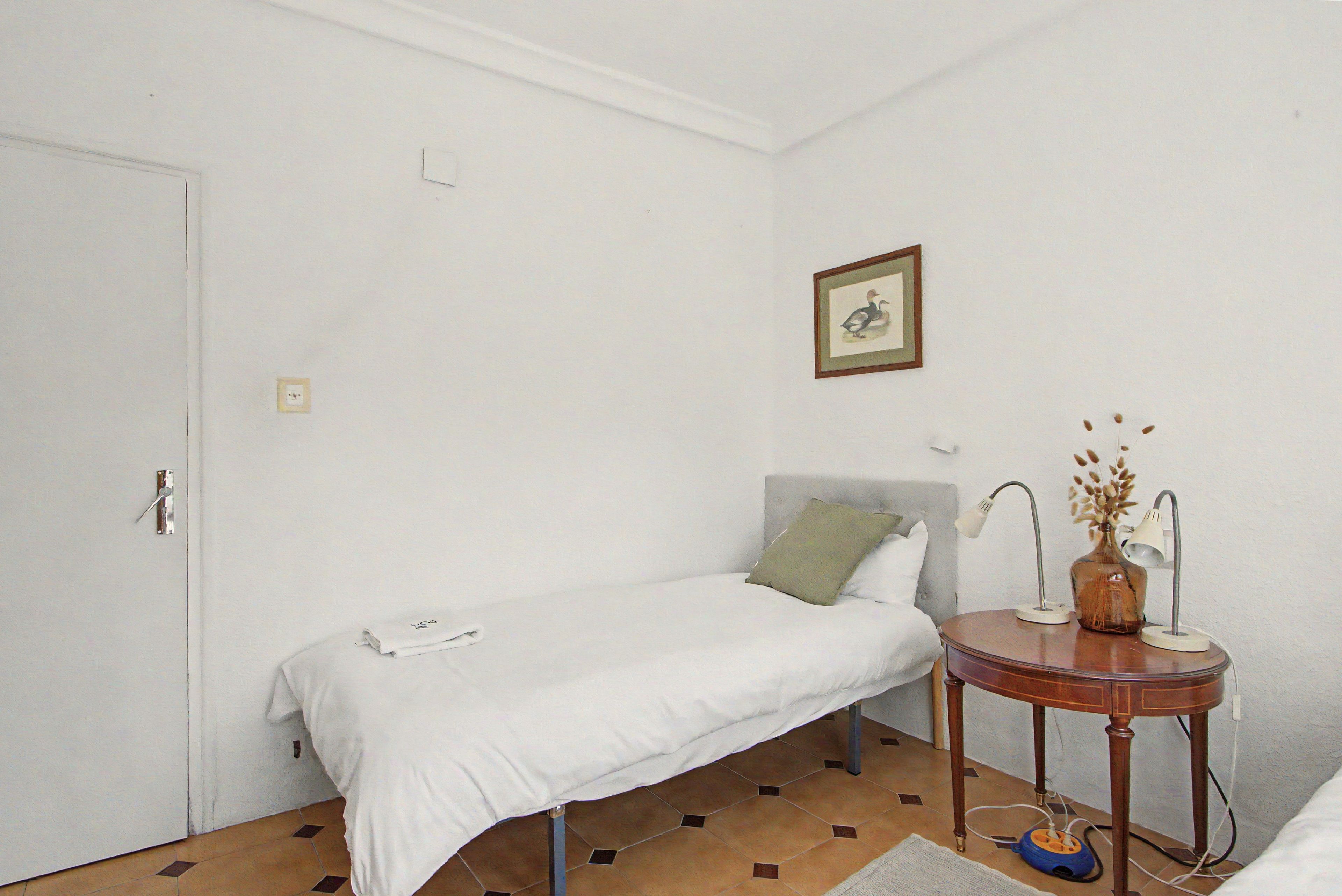 Una habitación pequeña y sencilla con una cama individual, una mesita de noche con una lámpara y un cuadro enmarcado en la pared.
