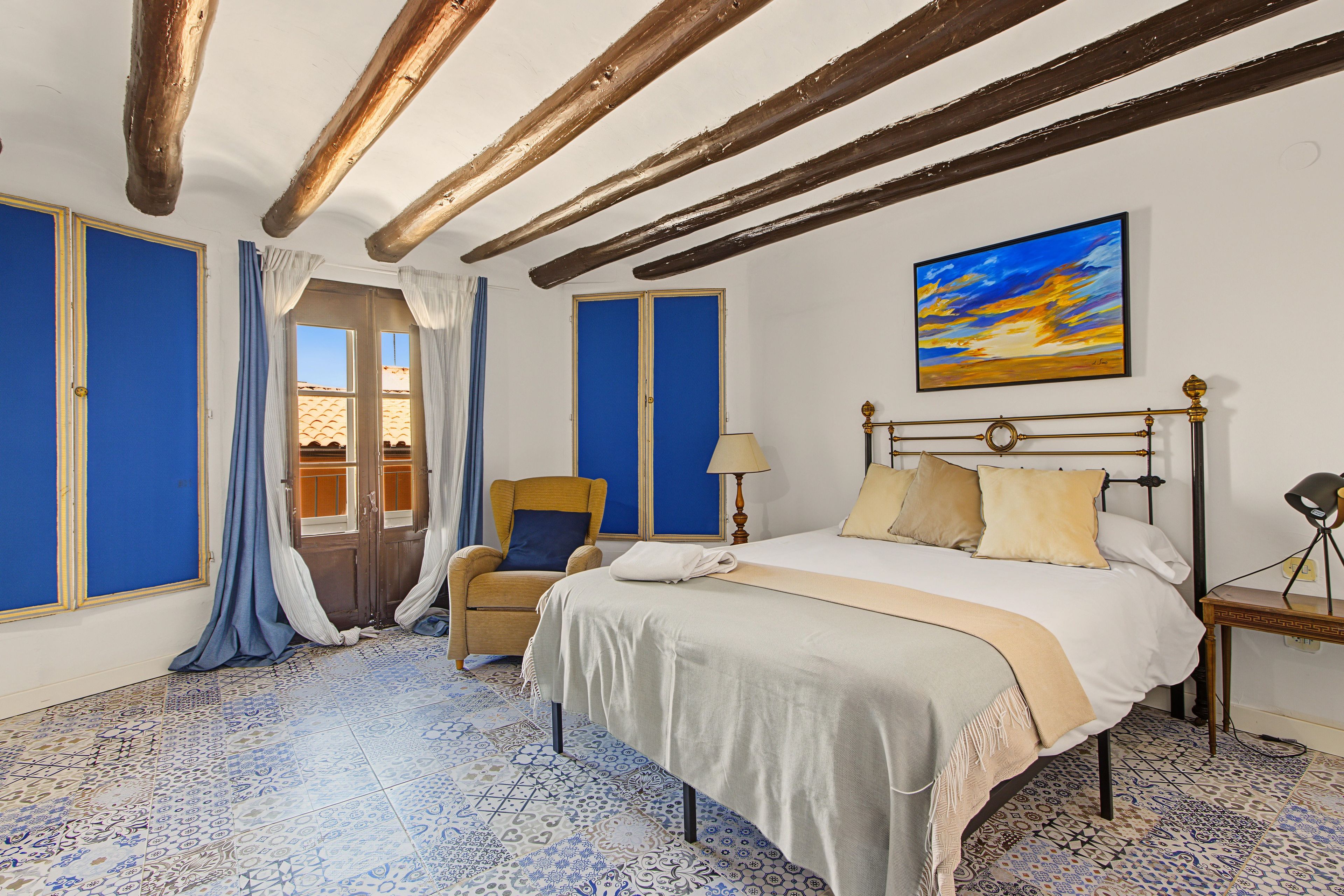A hotel room with a bed, a chair, a desk, and a painting on the wall.