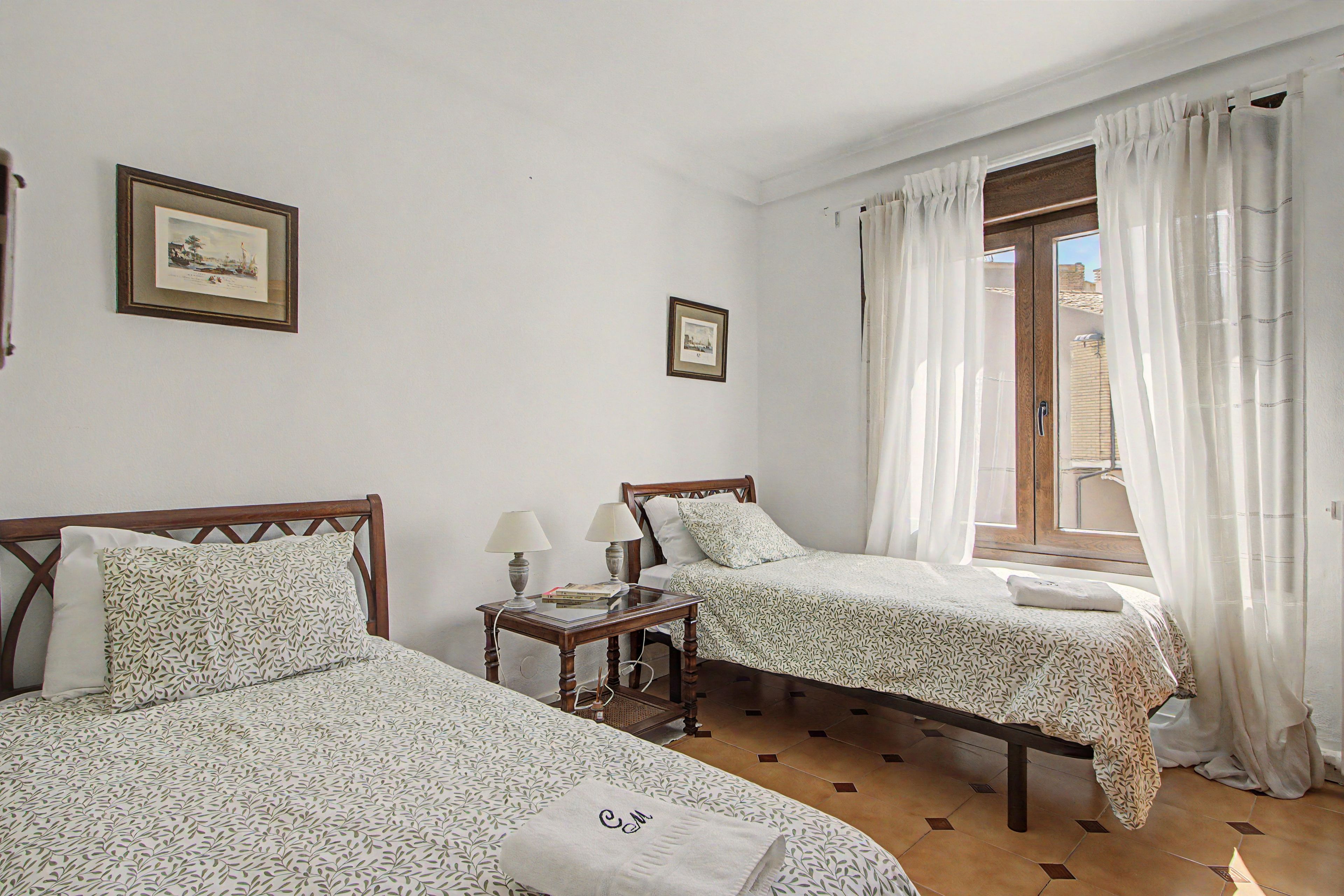 Una habitación con dos camas, una mesita con lámpara y un ventanal con cortinas.