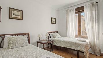 Una habitación con dos camas, una mesita con lámpara y un ventanal con cortinas.