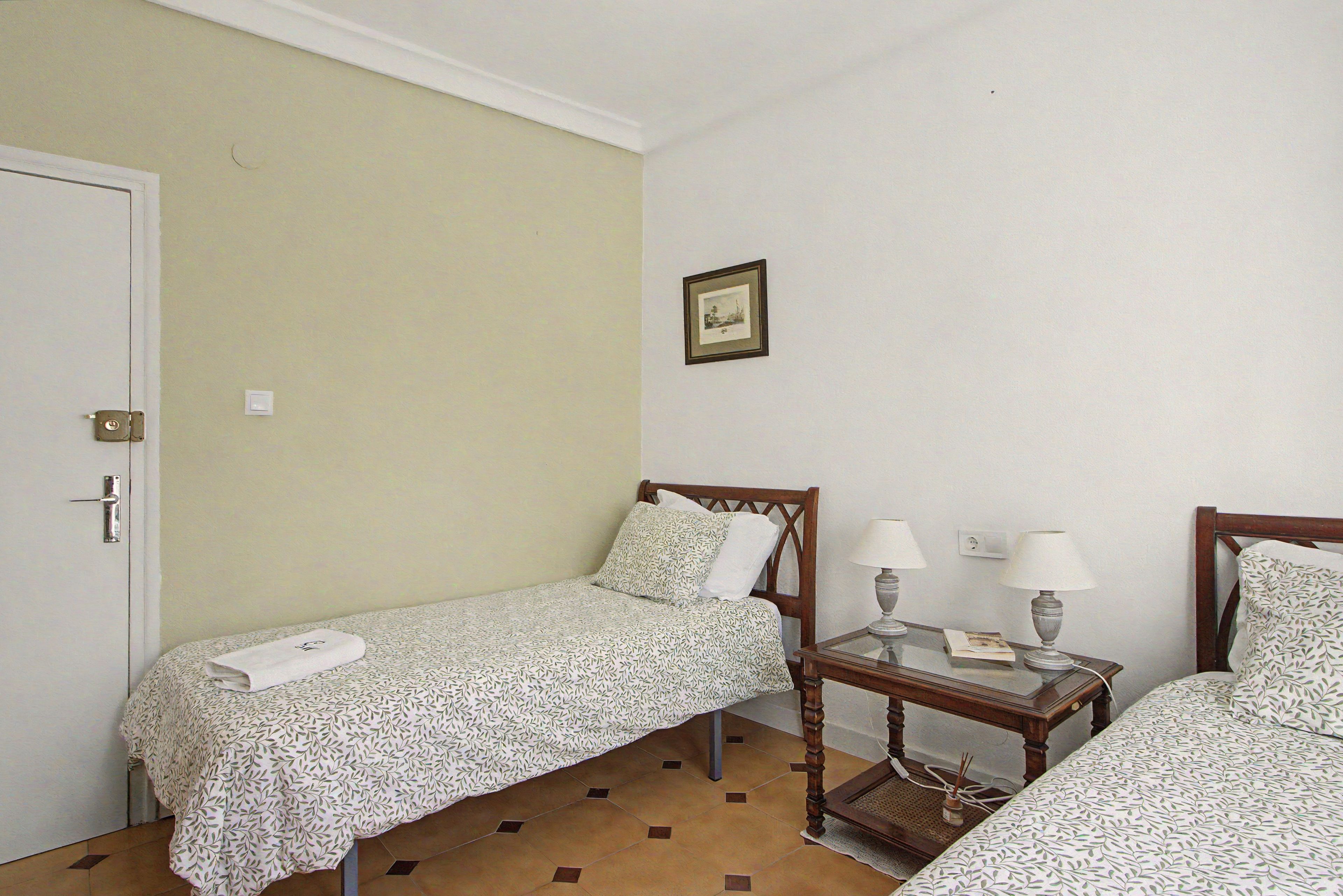 Una habitación sencilla con dos camas individuales, una mesita con dos lámparas y un cuadro enmarcado en la pared.