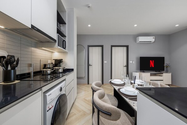 Une cuisine moderne équipée d’un lave-vaisselle, d’un four à micro-ondes et d’une cuisinière. On y trouve également un coin repas avec une table pour deux personnes et un téléviseur à écran plat affichant le logo de Netflix.