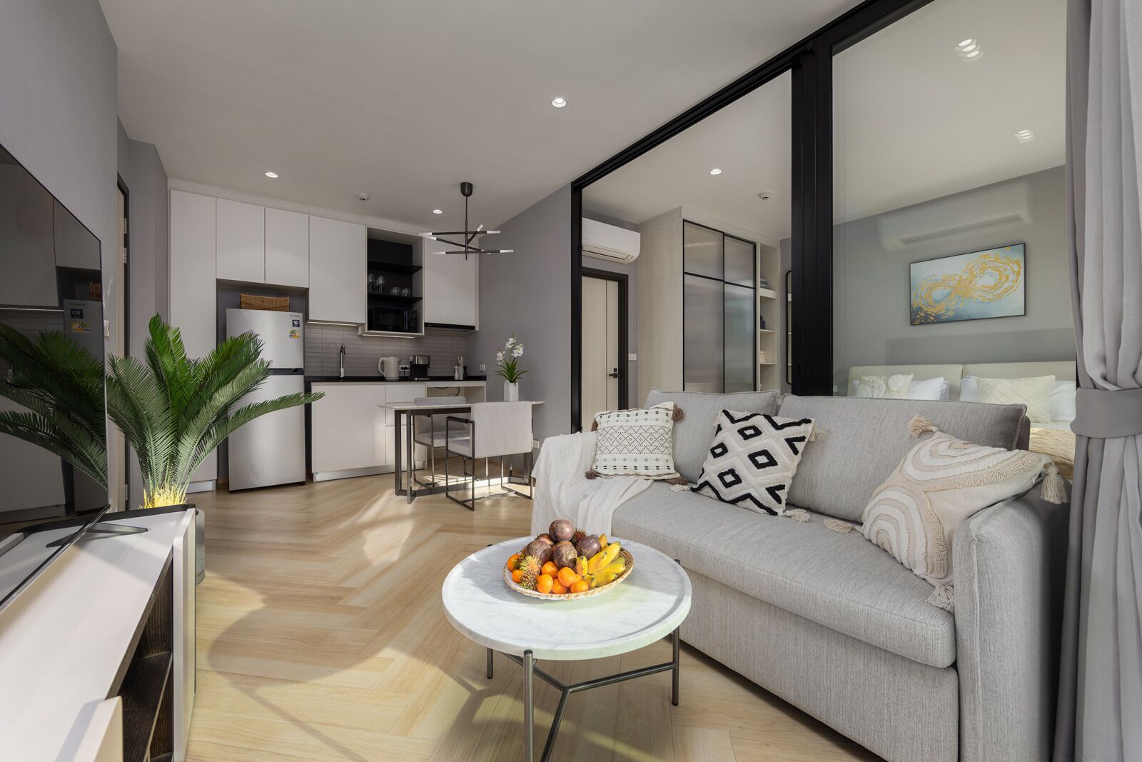 Una sala de estar moderna con un sofá, una mesa de centro redonda con fruta, una cocina al fondo y un ventanal grande.