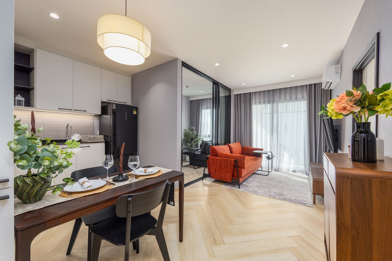 A modern living room with a sofa, a dining area with a table set for two, and a kitchen with a refrigerator.