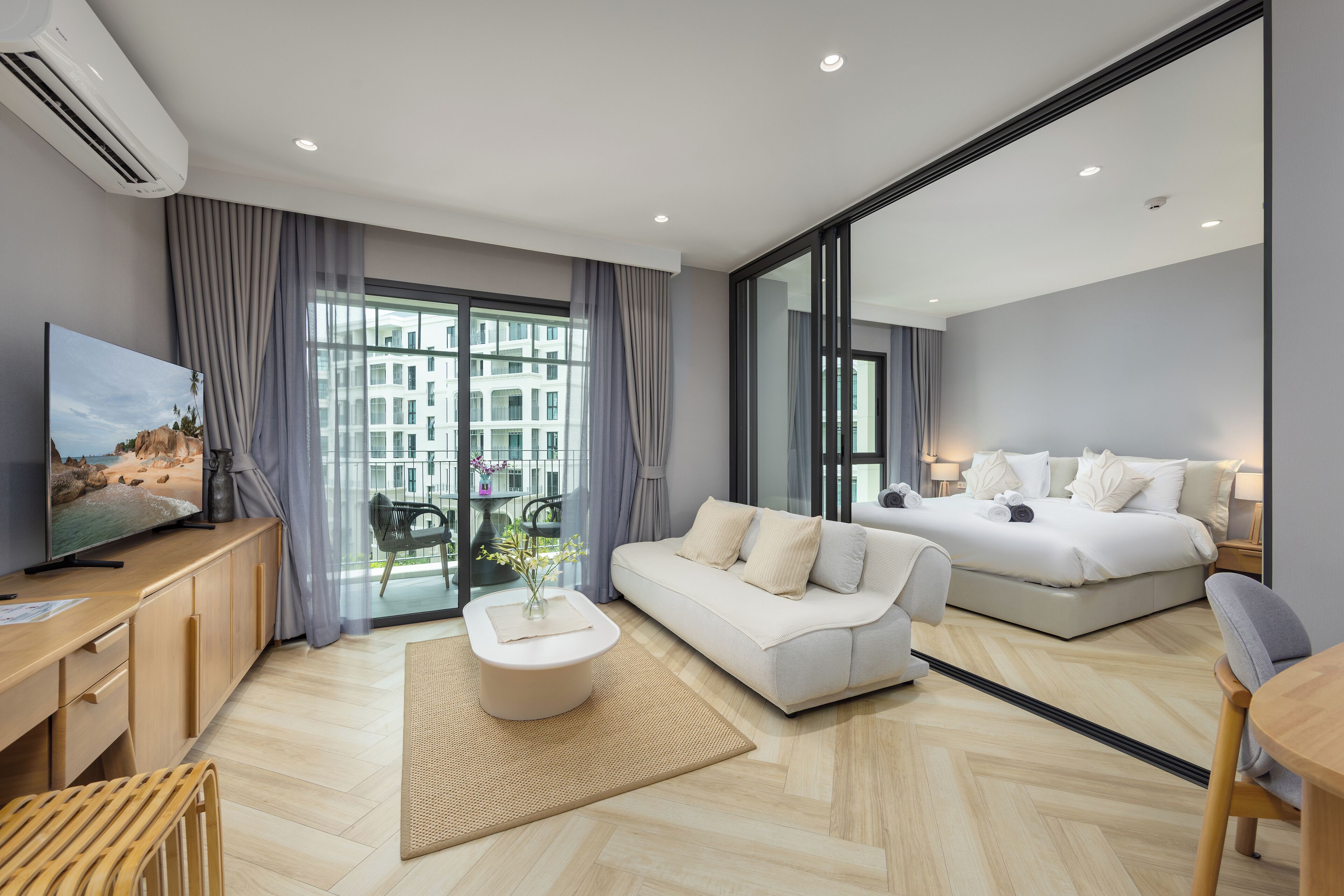 A modern hotel room with a sofa, a bed, a TV, and a balcony with a view of buildings.