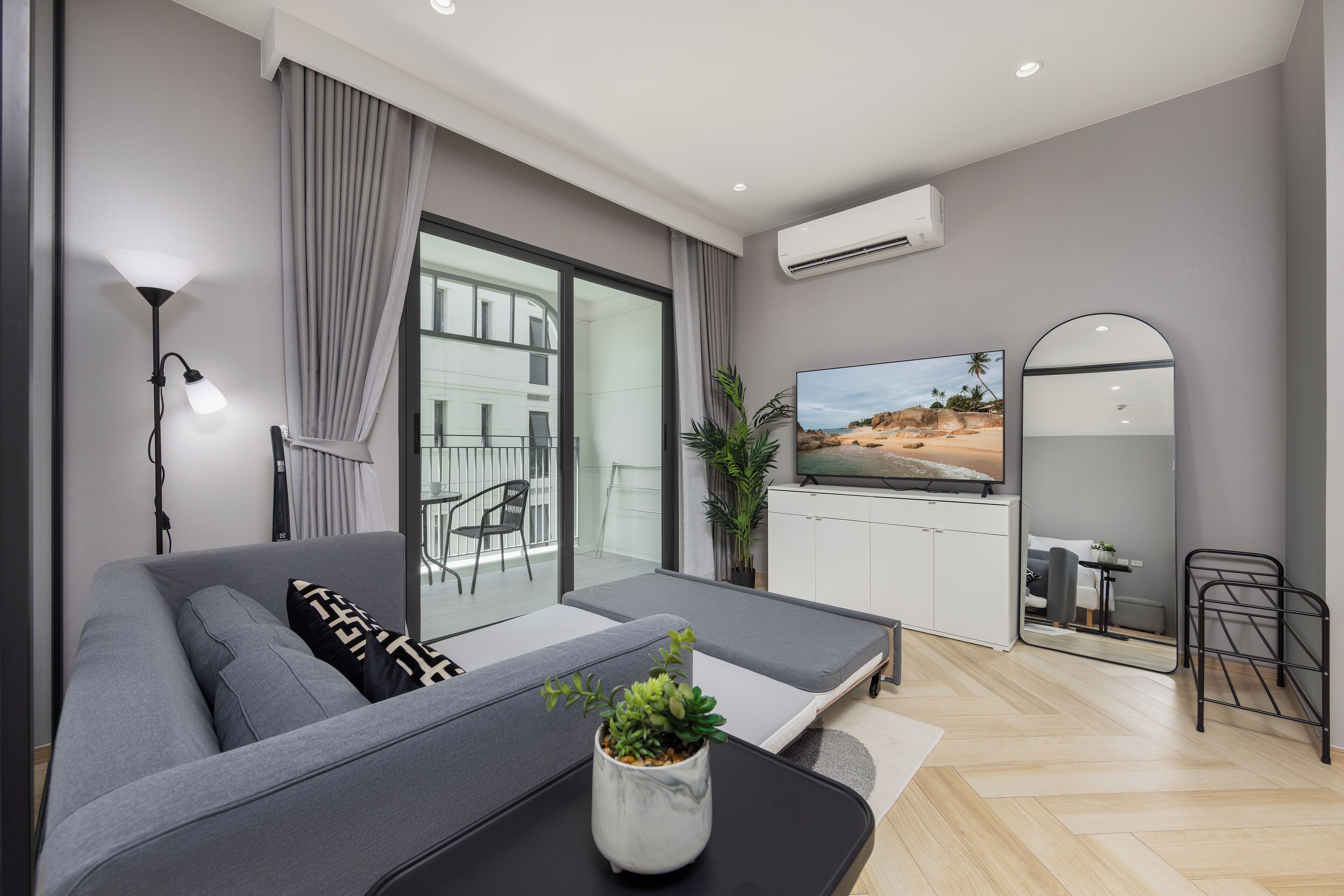 A modern living room with a grey sofa, a small table with a plant, a TV, and a large window with curtains.