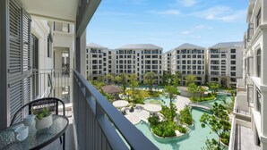 A balcony with a table, chairs, and a potted plant. In the background, there is a swimming pool with a surrounding garden and multiple hotel buildings. - The Title Legendary BangTao by TropicLook (Choeng Thale)