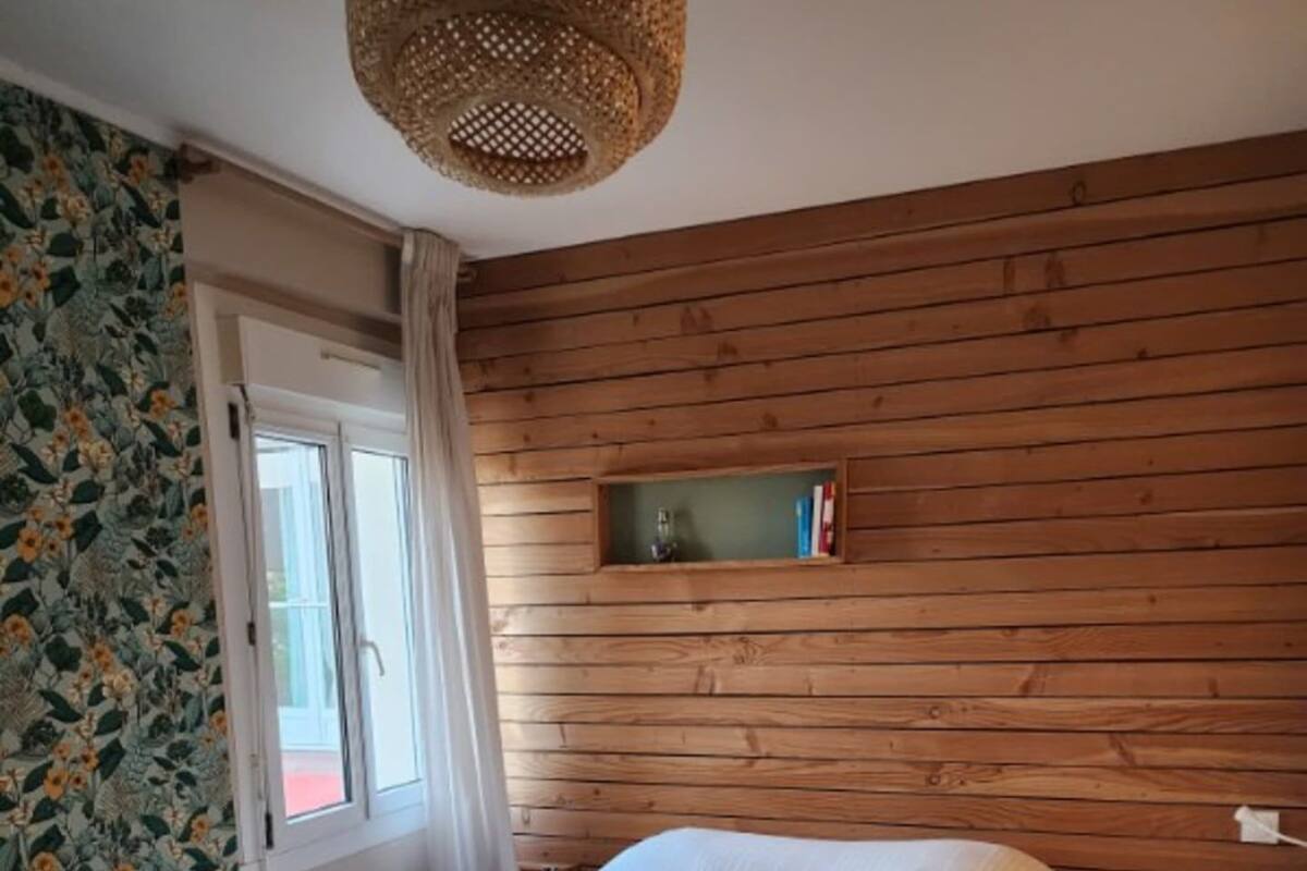 A bedroom with a wooden wall, a window with curtains, a bed with a white duvet, a small shelf with books, and a woven light fixture.