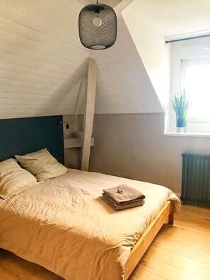 Ein Schlafzimmer mit einem hölzernen Bett, einer Pendelleuchte, einem Fenster mit Pflanze und einer Heizung.