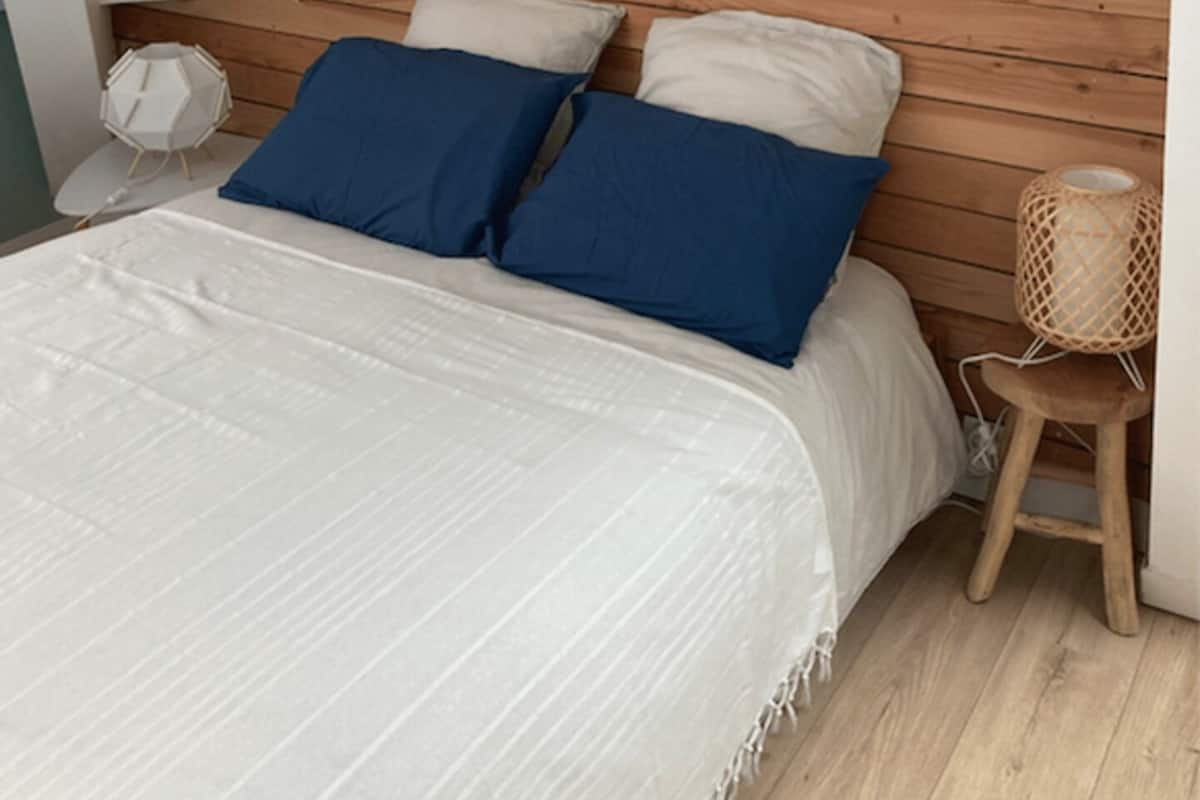 A bedroom with a wooden headboard, a bed with white bedding and blue pillows, a bedside lamp, and a small wooden stool with a woven light fixture.