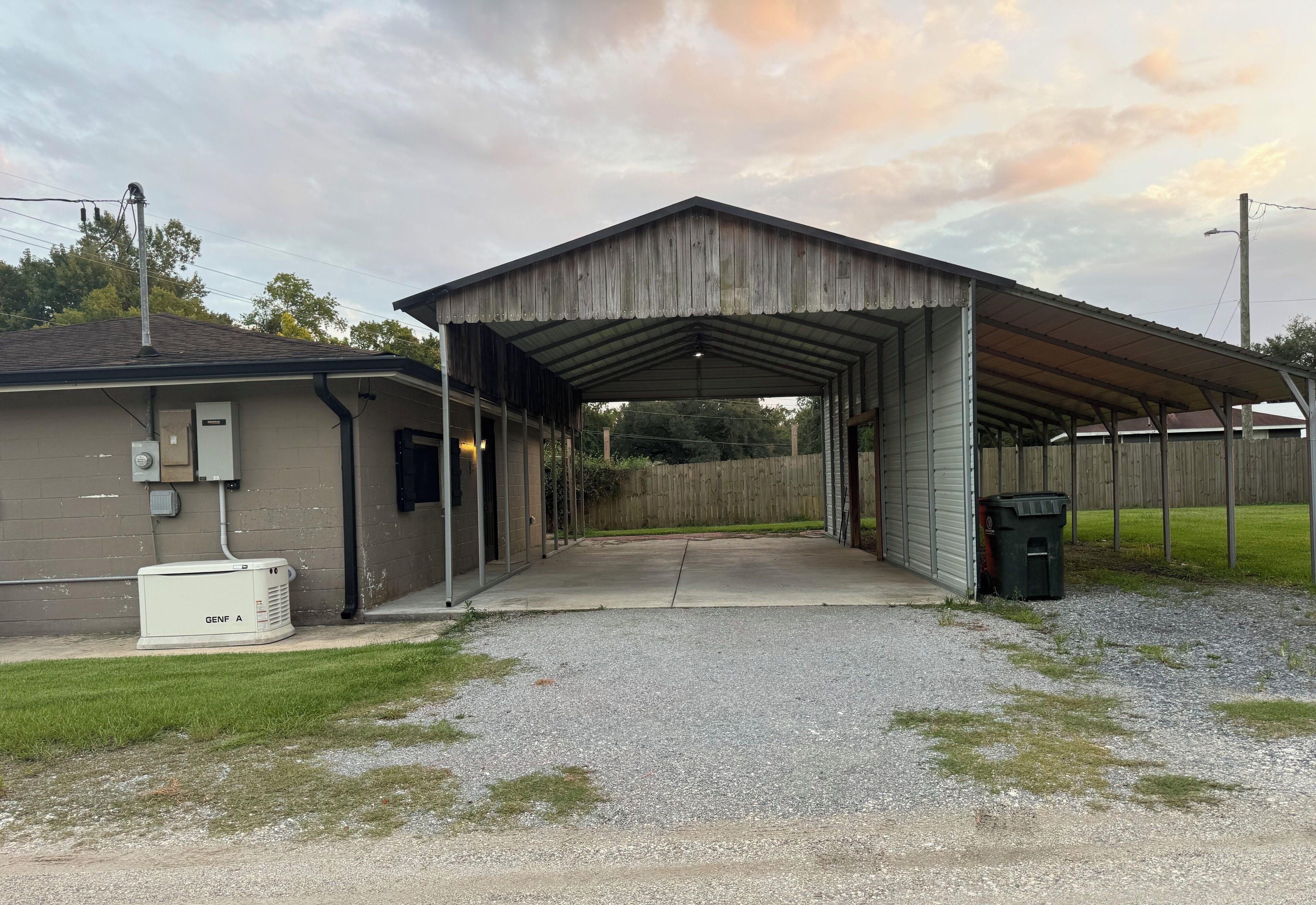 Un garage fermé avec une allée en gravier, un coffret électrique portant l’inscription « GENF A », et une clôture en bois en arrière-plan.