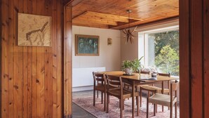 Un comedor de madera con una mesa, sillas, una lámpara colgante y un ventanal con vista a árboles.