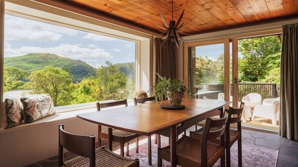 Un comedor con mesa y sillas de madera, ventanas amplias que ofrecen vistas a colinas verdes y un ventilador de techo.