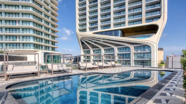 A modern hotel with a curved pool area, lounge chairs, and a view of adjacent high-rise buildings. - The Official Mercury Suite With Pool & Strong WIFI (St. Julian`s)