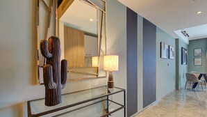 A modern hotel lobby with a large mirror, a cactus sculpture, a glass console table with a lamp, and a dining area in the background. - The Official Mercury Suite With Pool & Strong WIFI (St. Julian`s)