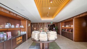 A hotel lobby with a central round table displaying towels, wooden shelving with bags, and a ceiling with a geometric pattern. - The Official Mercury Suite With Pool & Strong WIFI (St. Julian`s)