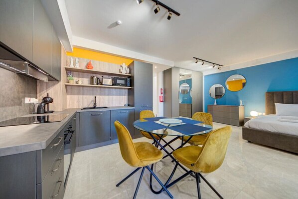 Una habitación de hotel moderna con cocina, un comedor con sillas amarillas, una cama con cabecera gris y una pared de acento azul.