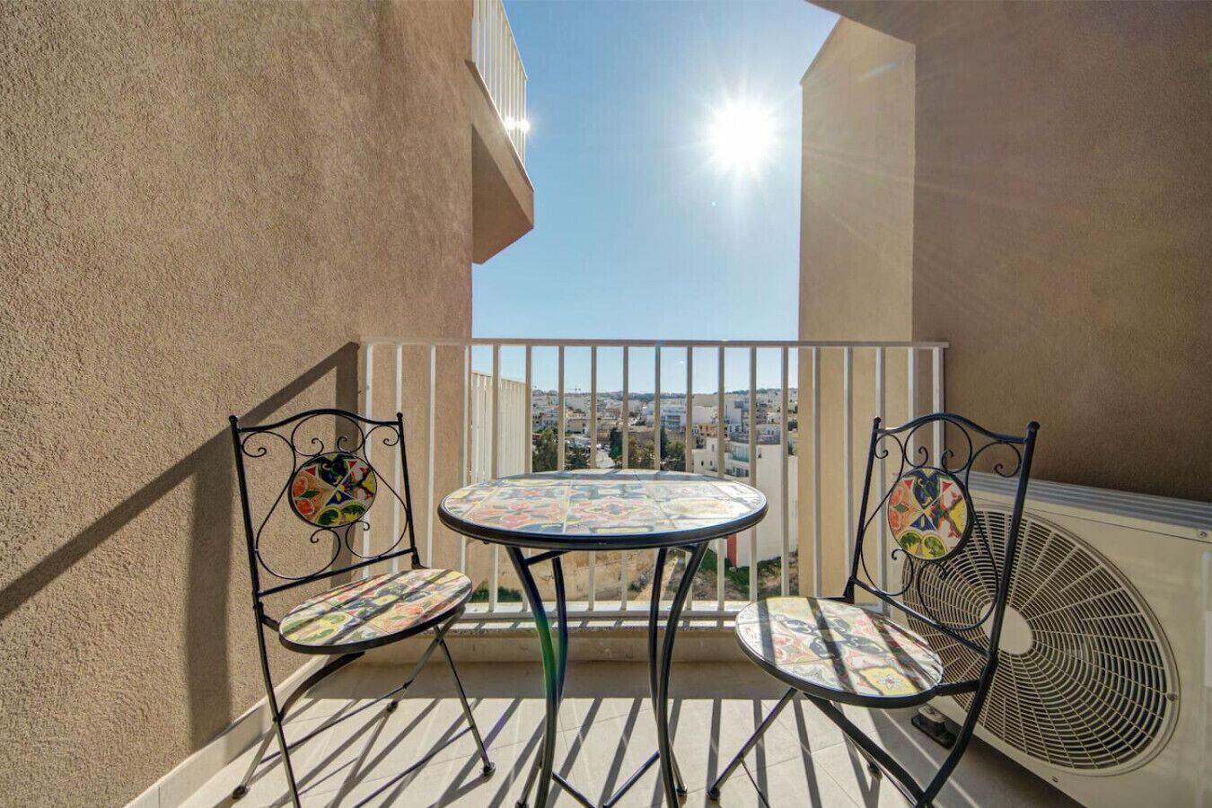 A balcony with a round table and two chairs, a white railing, and a view of a cityscape.