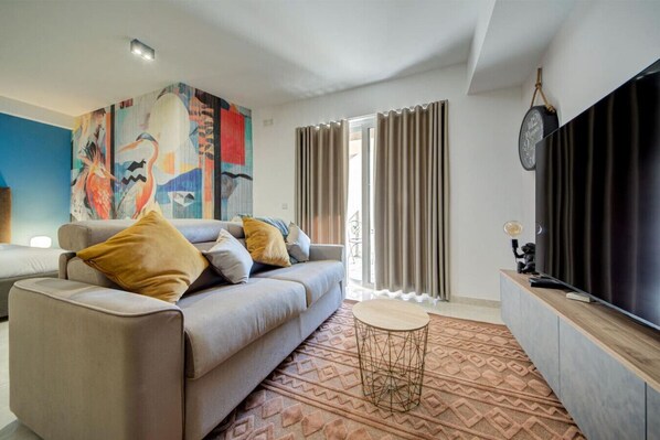 A modern living room with a sofa, a coffee table, and a large TV. There is a colorful abstract painting on the wall and a patterned rug on the floor.