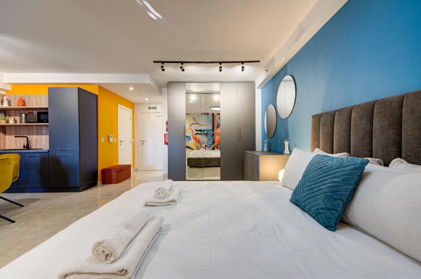 A modern hotel room with a large bed, a separate seating area with a yellow chair, a kitchenette, and a bathroom with a glass door.