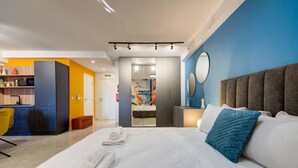 Une chambre d’hôtel moderne dotée d’un grand lit, d’un coin salon séparé avec un fauteuil jaune, d’une kitchenette et d’une salle de bain avec une porte en verre.