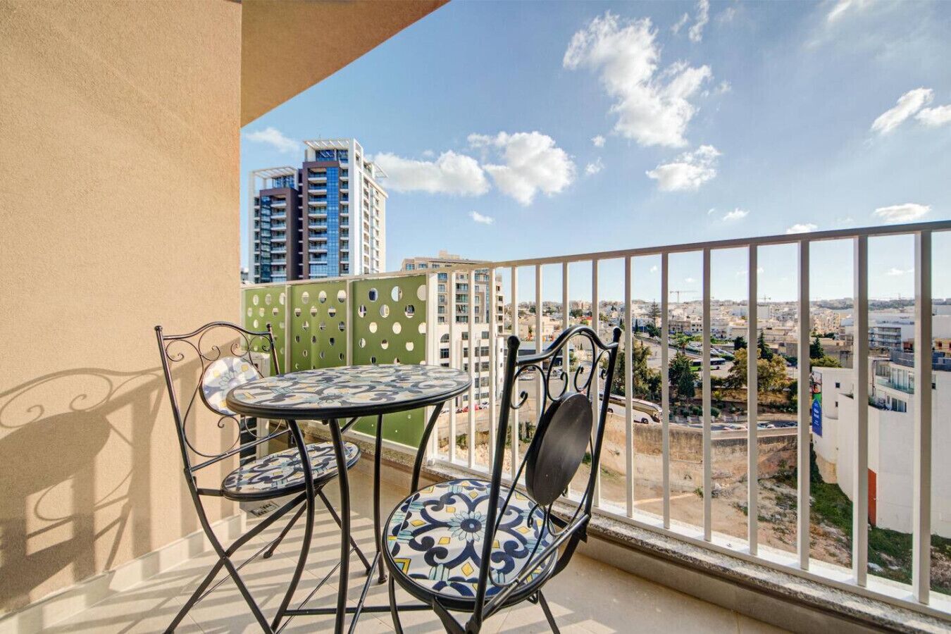 A balcony with a table and chairs, offering a view of a cityscape with buildings and a clear sky.