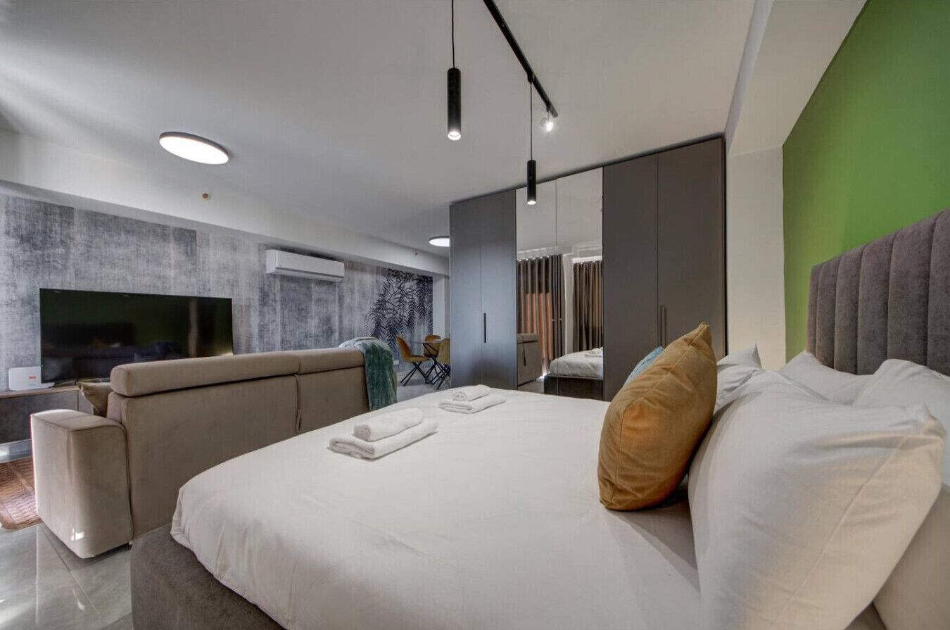 A modern hotel room with a large bed, a sofa, a flat-screen TV, and a green accent wall.