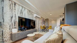 A modern living room with a large flat-screen TV, a sofa, a dining area with yellow chairs, and a patterned wallpaper. - Modern City Home | Walk to Nightlife & Dining (St. Julian`s)