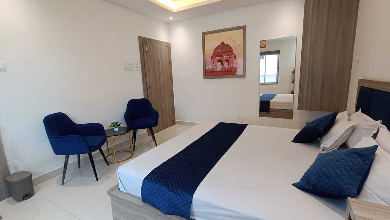 A hotel room with a large bed, two blue chairs, a small table, and a painting on the wall.